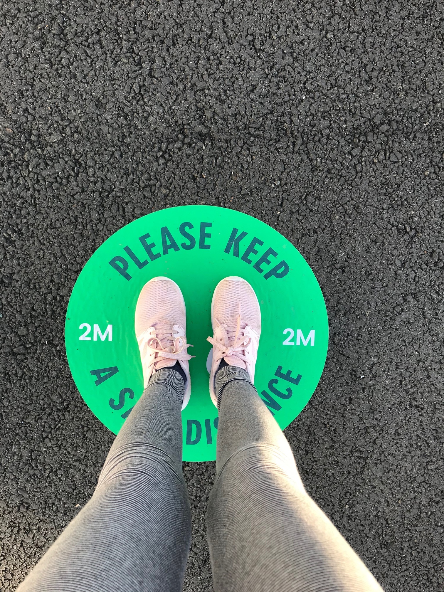 Photograph of someone's feet, standing on a green circle which says "Please Keep a Safe Distance"