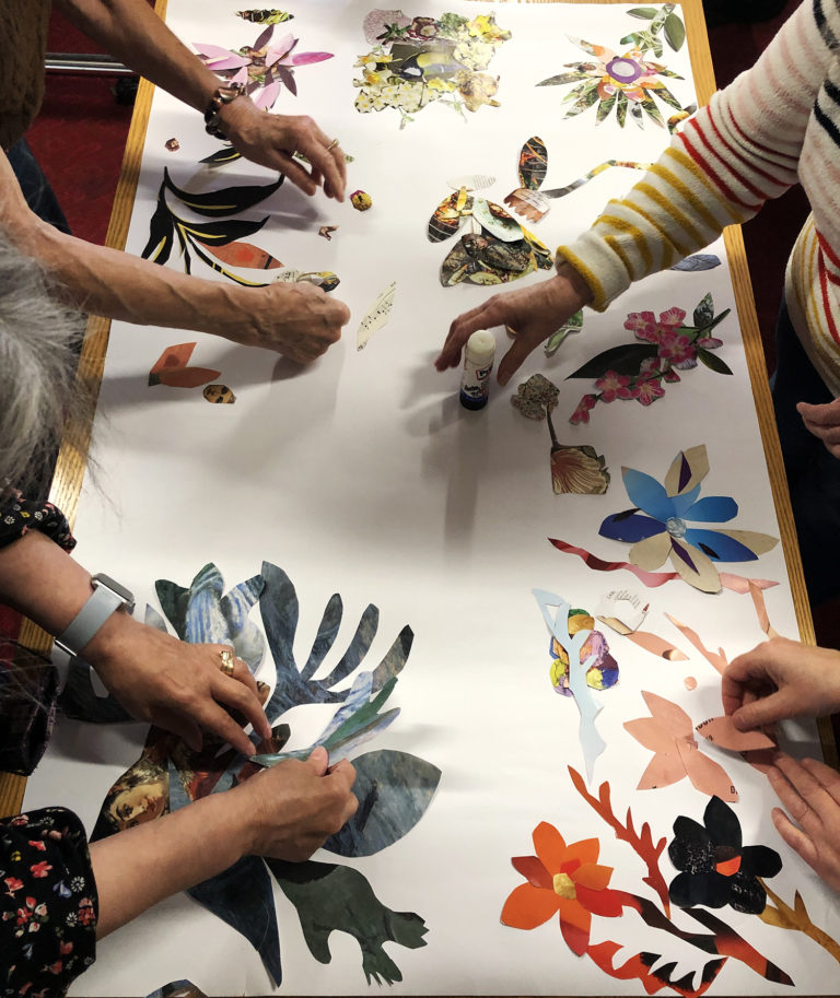 Hands of art makers from Garnethill Multicultural Centre's Senior Art Class, placing floral collage pieces on a large paper background.