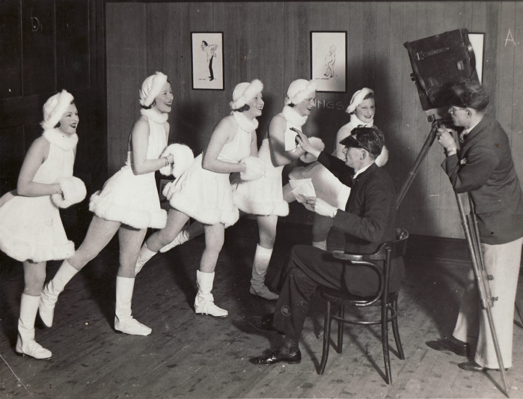 Black and white photograph showing 5 female dancers dressed in short white dresses with matching hats and muffs. All dancers standing as if ice-skating, in front of a director and cameraman, both men dressed in suit jacket and trousers.