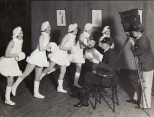 Black and white photogrpah showing 5 femal dancers dressed in short white dresses with matching hats and muffs. All dancers tandign as if ice-skating, in front of a director and cameraman, both men dressed in suit jacket and trousers.
