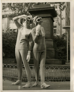 Black and white photo of two female models wearing woollen jumpers, leggings and boots, with woollen mittens, hats and goggles, standing in front of a statue on a plinth.