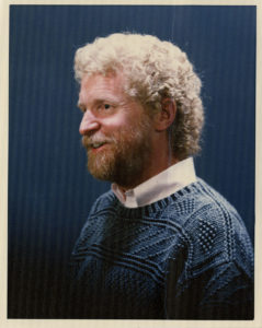 Portrait photograph of man with white curly hair and auburn beard, wearing a white shirt and blue woollen jumper