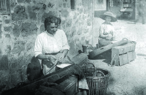 Black and white photo of two women seated, baiting lines, in front of a building and with bowls and buckets around them.