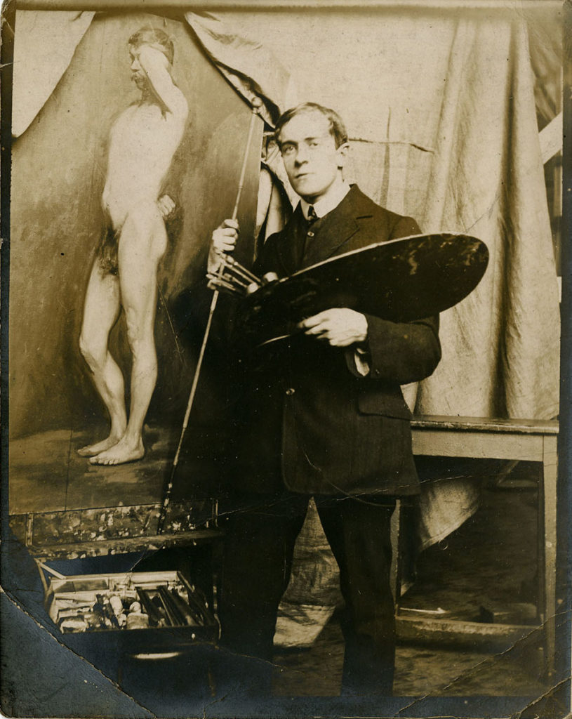 Archibald Haswell Miller poses with a palette and brushes in front of a painting of a male nude