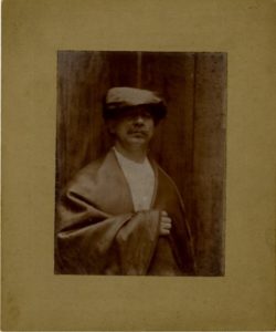 Sepia-coloured photograph of man wearing dark cloak and hat, with white shirt. Man has a moustache.